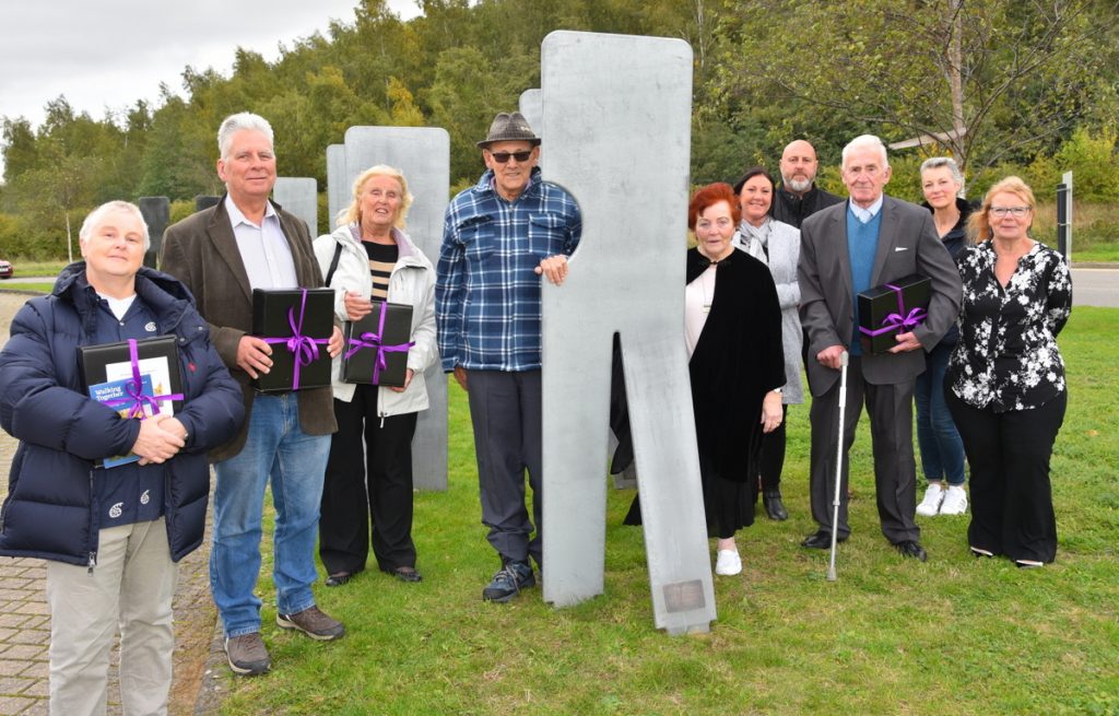 A group of about 11 people stand around life size abstract steel figures outside on grass.